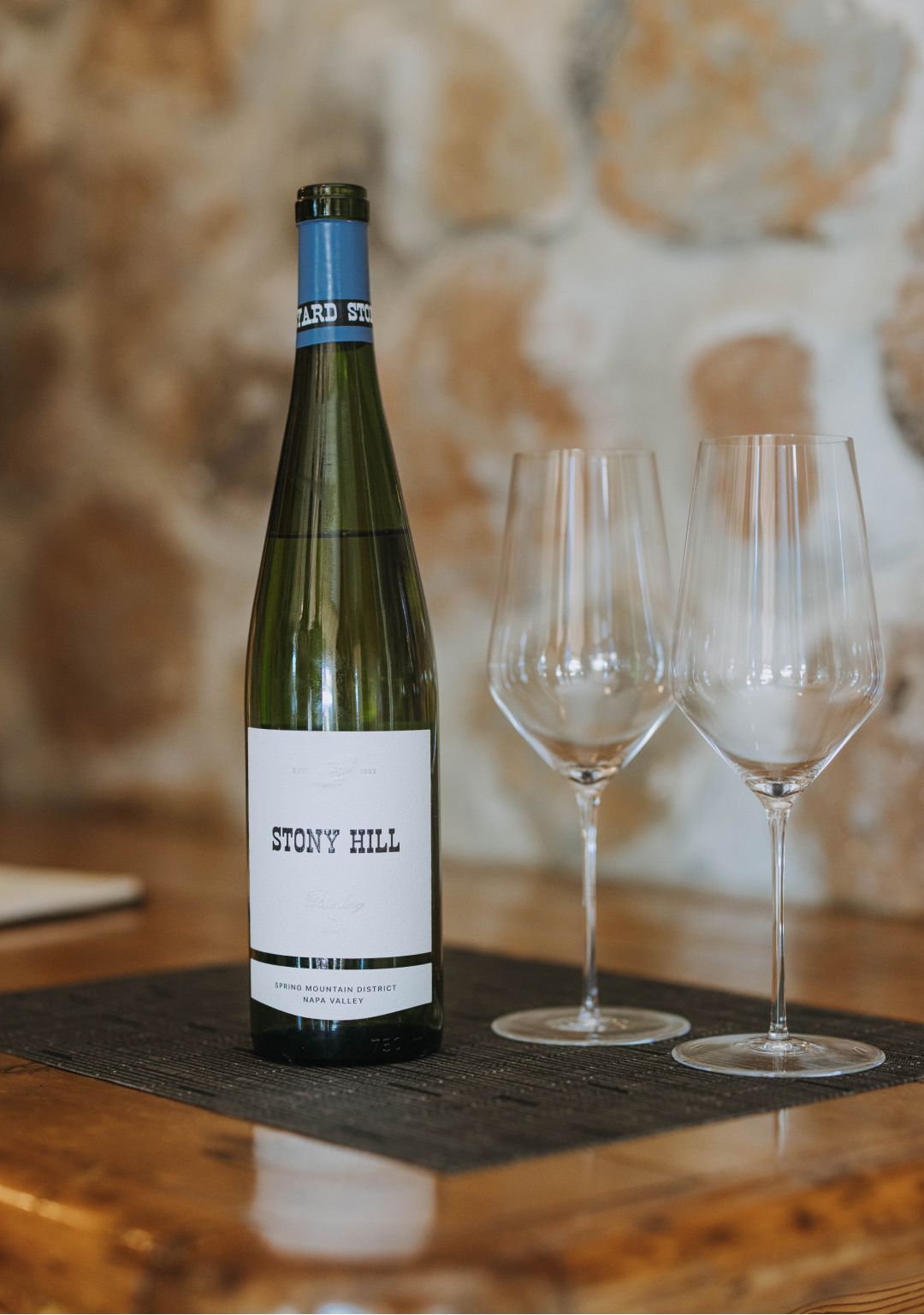 A bottle of Stony Hill wine with two glasses on a table in front op a stone wall at the Stony Hill residence.