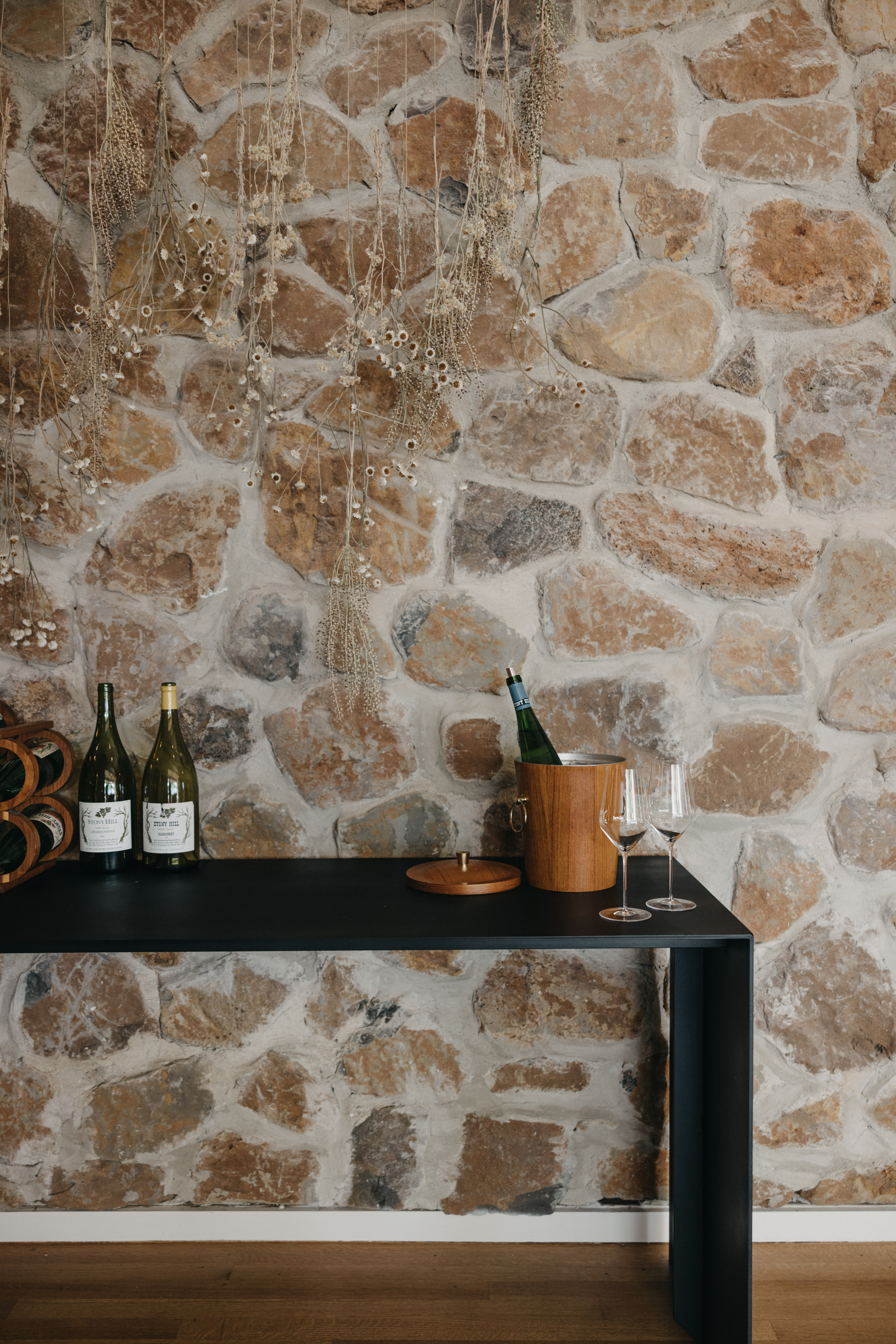 A stone wall with a table in front, two bottles of Stony Hill wines on the table, with another bottle of Stony Hill in a ice bucket and a glass of wine next to it.