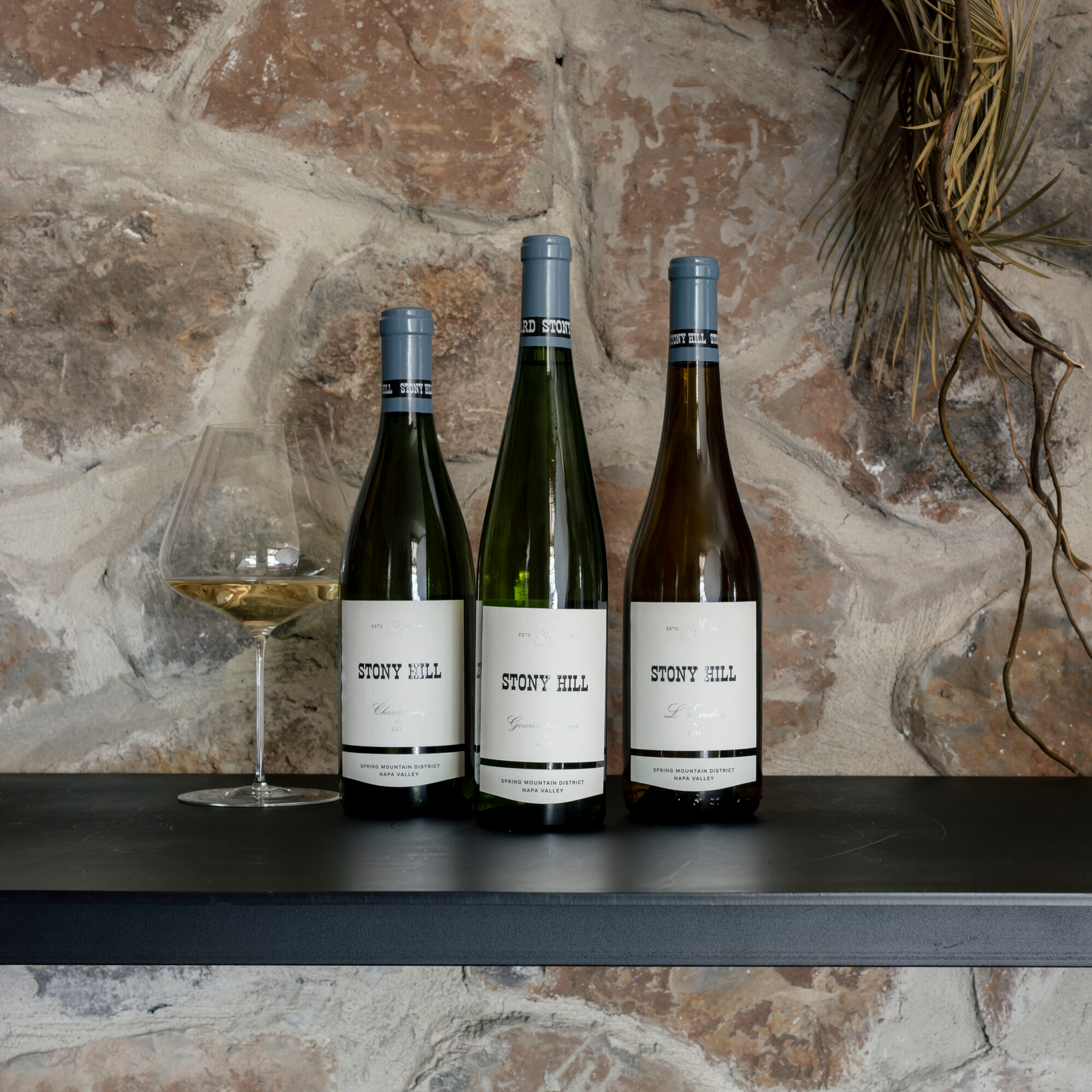 Stony Hill The Signature Trio: A bottle of 2021 L'Escalier, 2020 Chardonnay, and 2020 Gewurztraminer on a table next to a glass of wine.